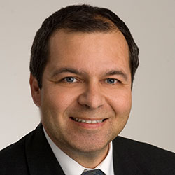Werner_Koch Ein Mann mit kurzen dunklen Haaren, der ein schwarzes Jackett, ein weißes Hemd und eine dunkle Krawatte trägt, lächelt vor einem schlichten hellen Hintergrund in die Kamera.