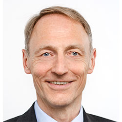 Wemhoener_Frederik_Dr_2022_0153_34 Ein Mann mittleren Alters mit heller Haut und kurzem hellbraunem Haar, der einen Anzug trägt und lächelt, posiert vor einem schlichten weißen Hintergrund.