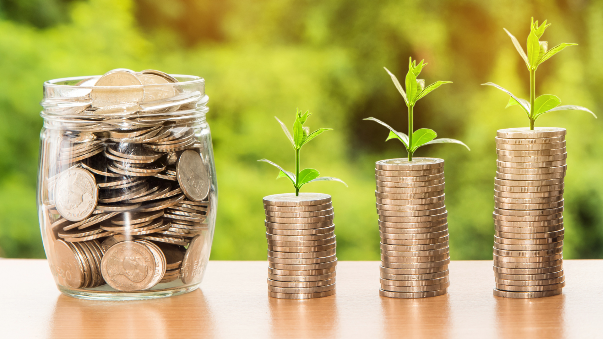 Neben einem mit Münzen gefüllten Glas stehen drei Münzstapel, auf denen jeweils eine kleine grüne Pflanze sitzt. Sie symbolisieren finanzielles Wachstum und Investitionen – perfekt, um die Vorteile eines Vermögenschecks zu veranschaulichen.