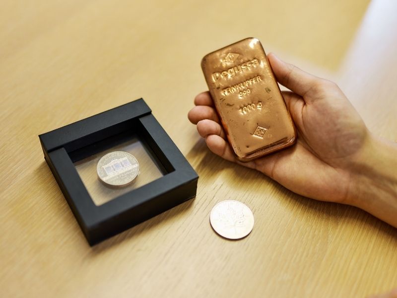 GFA FP_Edelmetalle Zwei Silbermünzen liegen auf einem Tisch, daneben ist eine Hand zu sehen, die einen Kupferbarren hält.