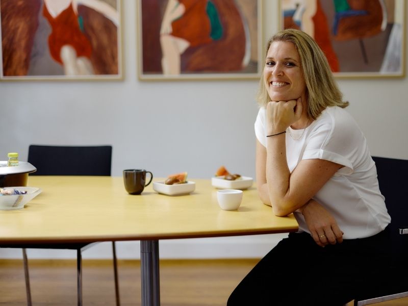 GFA FP_Frauen Finanzen Eine Frau sitzt am Tisch. Im Hintergrund sieht man Kunstgemälde.