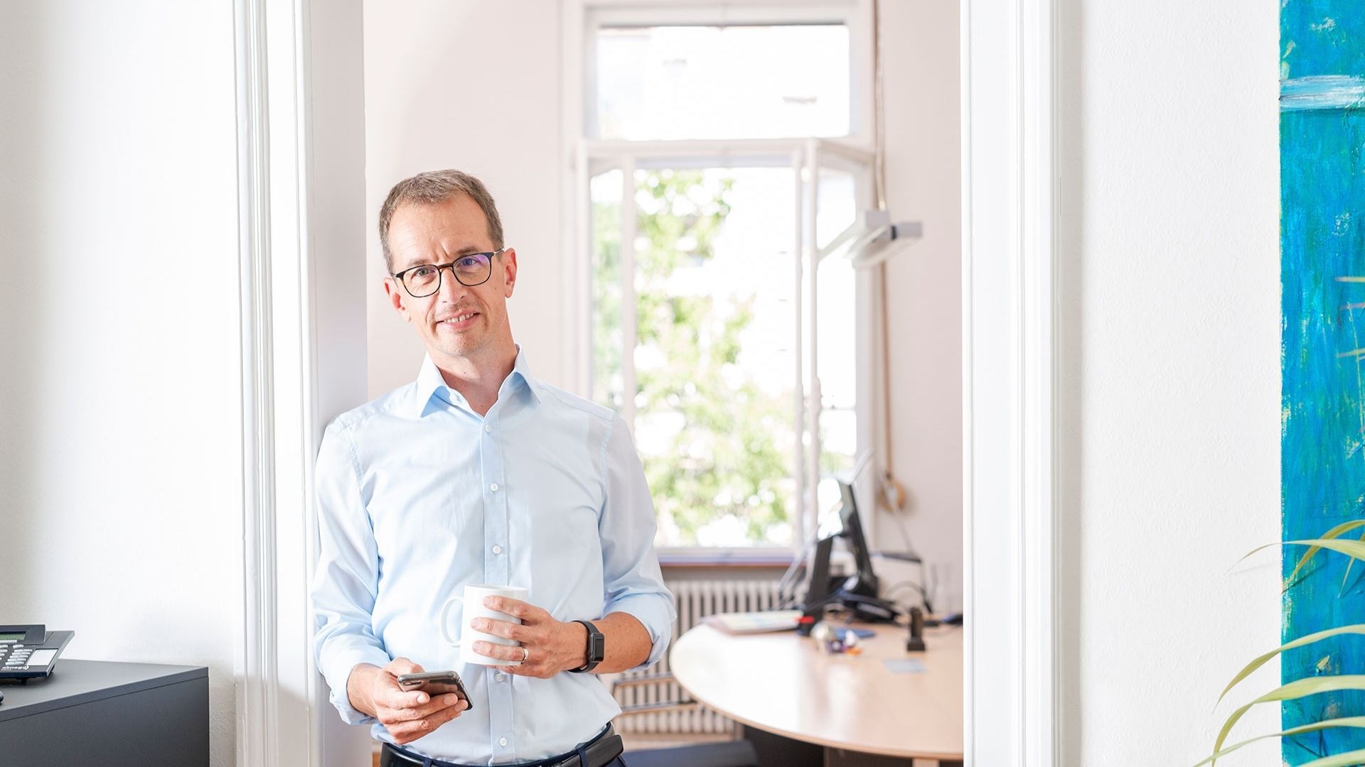 GFA FP_Header3 Ein Mann lehnt im Türrahmen mit Handy und Kaffeetasse in der Hand.