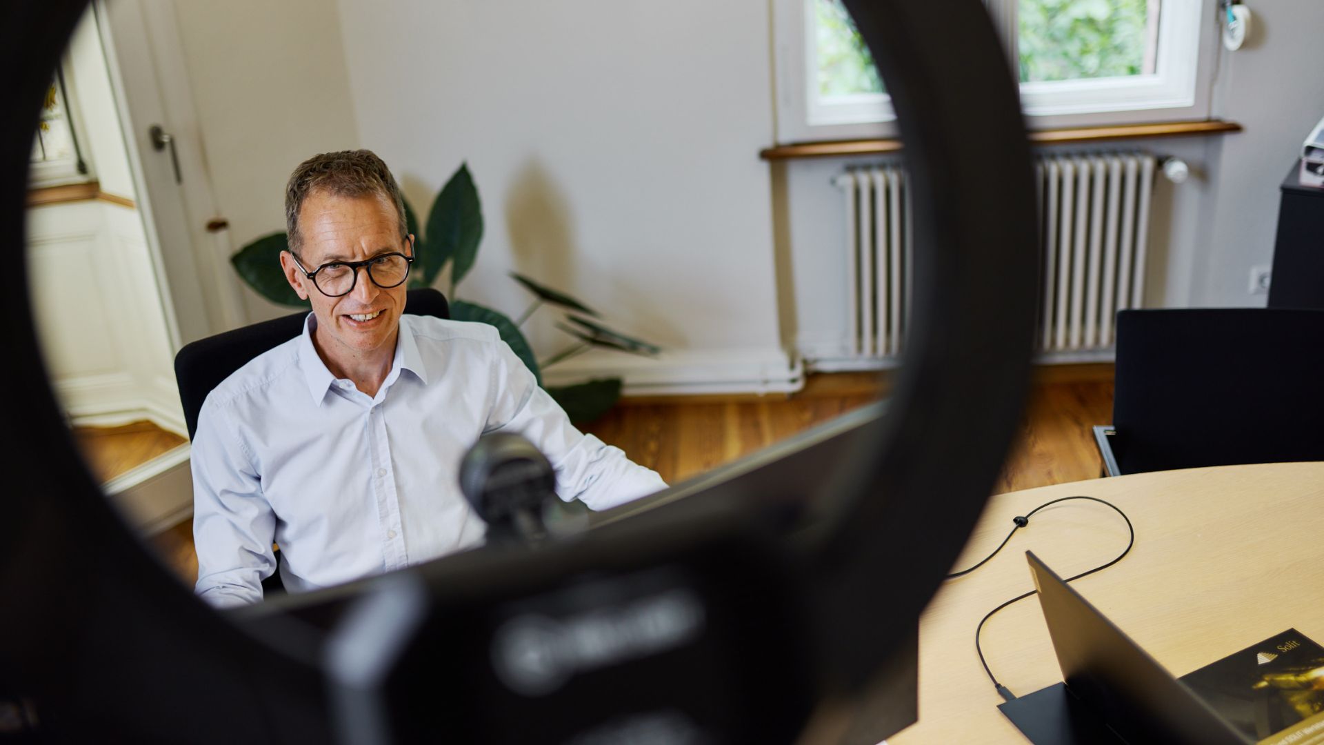 GFA VV_Videos Das Bild zeigt Mathias Lebtig durch ein Ringlicht fotografiert, der an seinem Schreibtisch sitzt und ein Video aufnimmt.