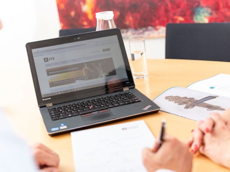 GFA FP_digitale VV EIn Laptop steht auf dem Tisch. Davor liegt ein Blatt Papier und eine Hand, die einen Kugelschreiber hält.